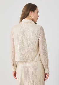 Jachetă bej din materiale cu paiete, cu mâneci lungi și guler, având dungi verticale din paiete reflectorizante și manșete cu nasturi.