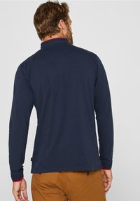 Mörkblå långärmad polo skjorta med röd krage och ärmslut, med en texturerad tygkvalitet och rak fåll. Bakifrån visas minimala designdetaljer.