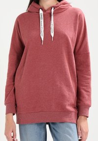 Ruskotoinen huppari valkoisilla nyöreillä, jossa lukee "VAIN koska se on intohimo," käytettynä vaaleansinisien farkkujen kanssa.