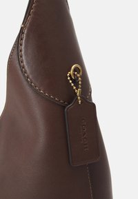 Coin de sac en cuir marron avec couture apparente et étiquette en cuir rectangulaire Coach New York attachée à une chaîne à billes dorée.