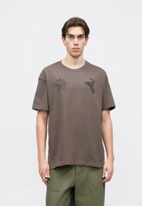 Καφέ βαμβακερό T-shirt με λαιμόκοψη και κοντά μανίκια. Διαθέτει μαύρα γραφικά στο στήθος, συμπεριλαμβανομένου του λογότυπου της μάρκας και vintage στοιχεία σχεδίασης.