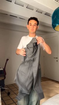 Jeune homme debout à l'intérieur tenant une veste grise, portant un t-shirt blanc et un jean bleu clair dans une pièce avec une lampe bleue et une chaise de bureau.