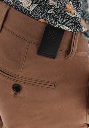 Brązowe spodnie o gładkiej fakturze, z tylną kieszenią zapinaną na guzik oraz czarną skórzaną metką z detalem logo. Kwiatowa koszula widoczna powyżej.