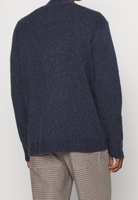 Tmavě modrý pletený svetr s texturovaným povrchem, s vysokým výstřihem a žebrovanými manžetami, doplněný o vzorované kalhoty.