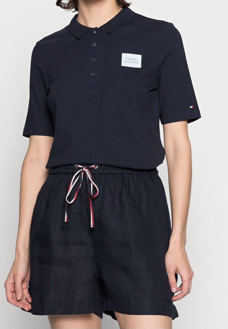 Personne portant un polo Tommy Hilfiger bleu marine avec patch logo et short assorti avec un cordon rouge, blanc et bleu marine à la taille.