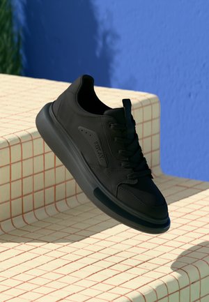 Sneaker nere con lacci, realizzate in pelle liscia, punta tonda, suola in gomma spessa e marchio discreto di lato, esposte su una superficie decorata.