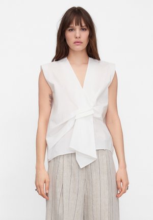 Młoda kobieta o prostych brązowych włosach, ubrana w białą, bezrękawną bluzkę typu wrap oraz jasne, szerokie spodnie w delikatne prążki, stojąca na białym tle.