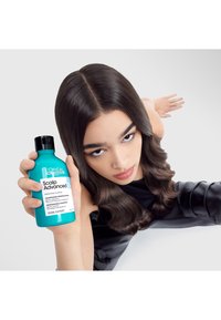 Turkos flaska med L'Oréal Scalp Advanced schampo, med svart kork och vit etikett, hålls av en person mot en ljusgrå bakgrund.