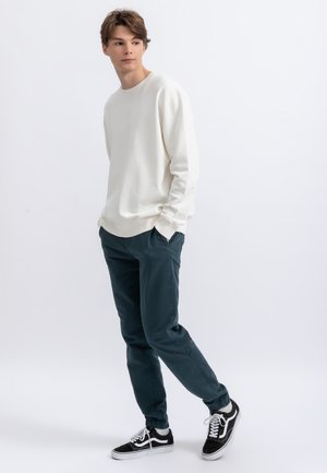 Crème-kleurige sweatshirt met lange mouwen, gecombineerd met donkerblauwe broek en zwarte sneakers met witte zolen. Casual, ontspannen pasvorm.