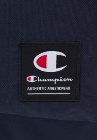 Patch en tissu noir avec un logo blanc et rouge comportant un "C" stylisé et le texte "Champion AUTHENTIC ATHLETICWEAR" sur fond sombre.
