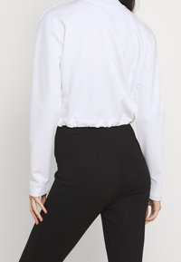 Sweatshirt blanc court avec des manches longues et une taille froncée, associé à un pantalon noir ajusté. Texture de tissu lisse et uni.