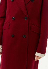 Burgundyröd ullblandad kappa med dubbelknäppning, svarta knappar och sidofickor. Har skräddarsydda slag och en mjuk textur.