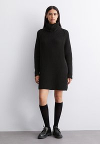 Svart turtleneckkjole laget av teksturert strikket stoff, med lange ermer og en avslappet passform, kombinert med knelengde sokker og svarte sko.