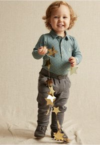 Sininen pitkähihainen poolopaita, jossa on pieniä pisteitä ja karhumerkki, harmaat tekstuuriset housut, lapsi pitää kädessään kultatähtiä, seisoo neutraalilla taustalla.