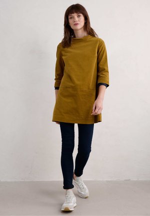 Mosterdkleurige tuniek met driekwartmouwen, uitgerust met een voorkantzak en een relaxte pasvorm, gecombineerd met donkere skinny jeans en witte sneakers.