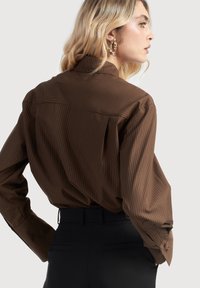 Hnedá pruhovaná blúzka s golierom, dlhými rukávmi a zapínačmi na manžetách, ktorá má hladkú textúru a voľný strih. Kombinovaná s čiernymi nohavicami.