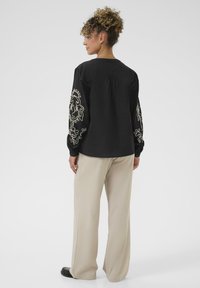 Zwarte blouse met geborduurde beige patronen op de mouwen, ontspannen fit, ronde halslijn, gecombineerd met lichtbeige wijde broek.
