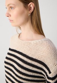 Strikkegenser i kremfarge med svarte striper. Har en bred utringning og et strukturert maskemønster. Utsmykket med en glatt overflate.