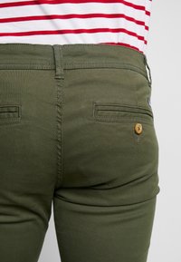 Olivgrüne Hose mit glatter Textur und schmaler Passform. Verfügt über zwei Gesäßtaschen und eine Holzknopf-Details.