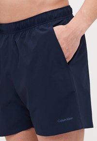 Námornícke modré plavky s elastickým pásom, bočnou vreckom a malým logom 'Calvin Klein' v svetlomodrej farbe na ľavej strane.