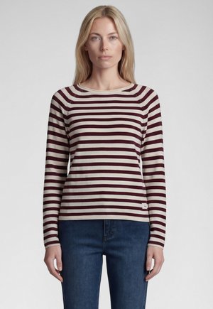 Woman with straight blonde hair wearing a long-sleeve cream and dark red striped shirt and blue jeans against a plain light background.