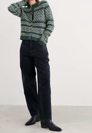 Personne portant un cardigan vert à motifs boutonné, un jean noir taille haute et des bottines noires, debout avec une main derrière la tête.