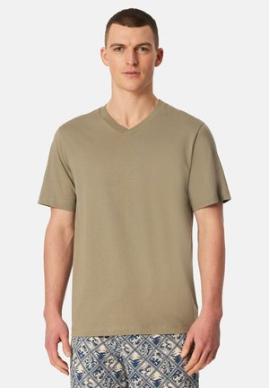 Mann steht und trägt ein schlichtes olivgrünes V-Ausschnitt-T-Shirt und blaue gemusterte Shorts vor einem weißen Hintergrund.