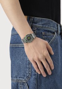 Poignet avec une montre en métal argenté dotée d'un cadran noir et vert, reposant près de la poche d'un jean en denim bleu et d'un haut noir.