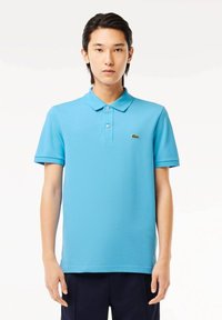 Polo de manga corta en tejido azul brillante, con un cuello clásico y un pequeño logo de cocodrilo verde en el lado izquierdo del pecho.