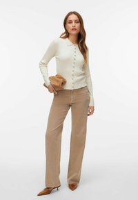 Żakiet kremowy prążkowany z kołnierzem i zapięciem na guziki, w połączeniu z beżowymi szerokimi spodniami i brązowymi szpilkami o szpiczastym nosku. Trzyma beżową kopertówkę.