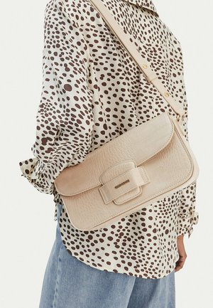 Mujer vestida con una blusa de lunares en crema y marrón y vaqueros azules, llevando un bolso bandolera de cuero texturizado beige con cierre de solapa.
