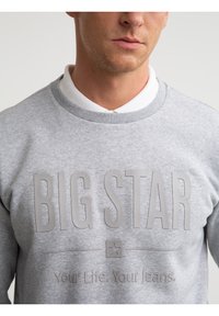 Mand iført lysegrå sweatshirt med "BIG STAR" og "Your Life. Your Jeans." præget på foran over en hvid skjorte med krave.
