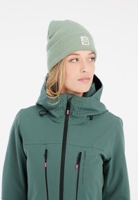 Grüne wasserdichte Jacke mit hohem Kragen, Reißverschluss vorne und Belüftungsöffnungen; kombiniert mit einer hellgrünen Mütze mit Logopatch.