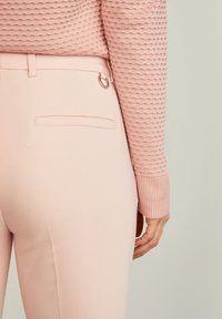 Pantaloni rosa chiaro con una texture liscia, dotati di una tasca posteriore e un dettaglio in metallo a forma di anello. Chi indossa tiene la vita con una mano.