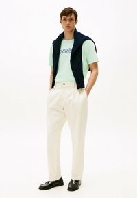 Camiseta de manga corta verde claro con texto azul, suéter anudado azul marino, pantalones crema y mocasines negros, todo sobre un fondo blanco.