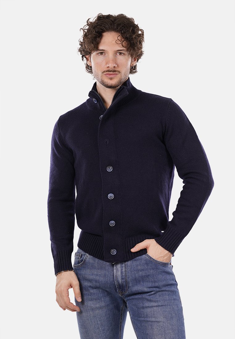 Cardigan lavorato a maglia di un blu navy scuro con colletto alto, sei bottoni, polsini e orlo a coste. Indossato sopra una camicia chiara e jeans aderenti.