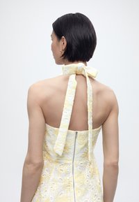 Γυναίκα με κοντά μαύρα μαλλιά φορώντας ένα κίτρινο και άσπρο φόρεμα με λουλούδια χωρίς τιράντες, δεμένο με φιόγκο στο πίσω μέρος του λαιμού, γυρισμένη με την πλάτη.