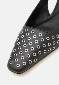 Schwarze Lederschuhe mit spitzer Zehe und ausgeschnittenen Seiten, verziert mit silbernen Ösen in einem gemusterten Arrangement über dem Oberteil.