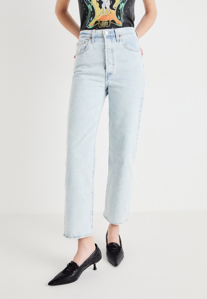 Personne portant un jean bleu clair taille haute à jambes droites et des chaussures noires à talons bas pointues, debout les mains dans le dos.