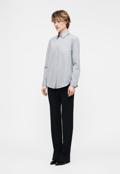 Άτομο που στέκεται φορώντας ανοιχτόγαλανη πουκαμίσα με κουμπιά, μαύρο παντελόνι με φαρδιά πόδια και μαύρα παπούτσια, σε απλό λευκό φόντο.