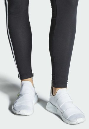 Zapatillas deportivas blancas con un diseño de tipo slip-on, que presentan correas cruzadas y tela texturizada. Combinadas con leggings negros con rayas laterales blancas.