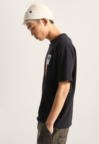 Sort kortærmet t-shirt med grafisk design på brystet. Båret med en lysfarvet ribbestrikket beanie og grå bukser, hvilket viser en afslappet pasform.