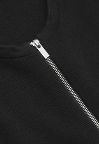 Svart stickad plagg med rund halsringning och detalj av silvrig metallzipper. Slät textur; zippen har metalltänder och ett polerat drag.