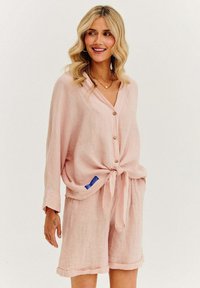 Bledě růžová lněná košile s designem na zavázání vpředu, dlouhými rukávy a dřevěnými knoflíky, v kombinaci se shodnými šortkami s ohrnutými lemy.