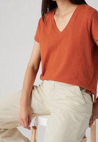 T-shirt V à col en rust orange en tissu doux, avec des manches courtes et une coupe décontractée, associé à un pantalon beige clair.