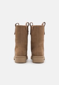 Bottines en suédine brune avec une texture lisse, des fermetures éclair latérales et des tirants en boucle. Semelles en caoutchouc épais couleur tan avec des rainures horizontales.