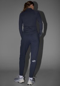 Femme aux cheveux bouclés en queue de cheval, vêtue d'un survêtement de sport foncé et de baskets grises, debout les mains dans les poches sur un fond gris.