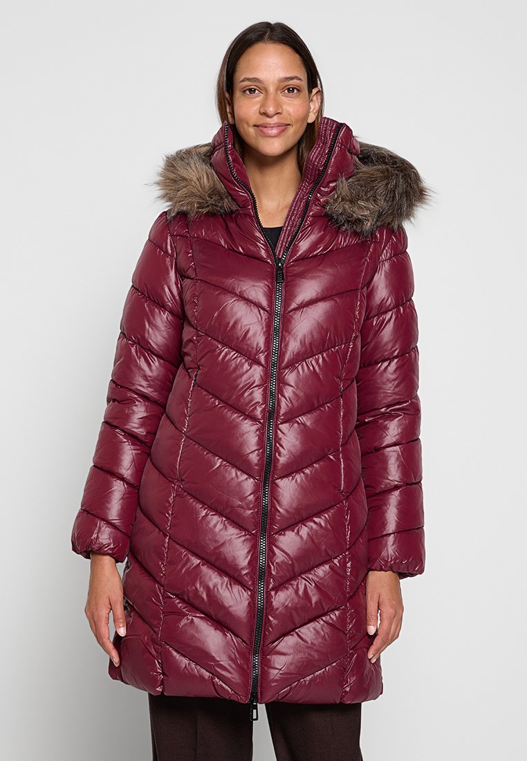 Vrouw in een lange, glanzende bordeauxrode gewatteerde pufferjas met een bontomzoomde capuchon, staande tegen een effen lichte achtergrond.