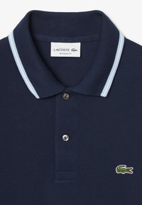 Polo de color azul marino hecho de material texturizado, con un cuello bicolor de azul claro, tapeta de dos botones y logo bordado de cocodrilo verde.
