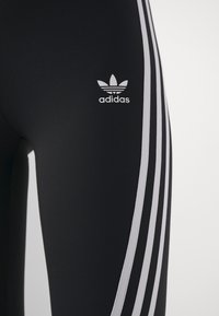 Svarta stretchiga leggings med tre vita ränder längs sidorna och en liten vit Adidas-logotyp på den övre låret.